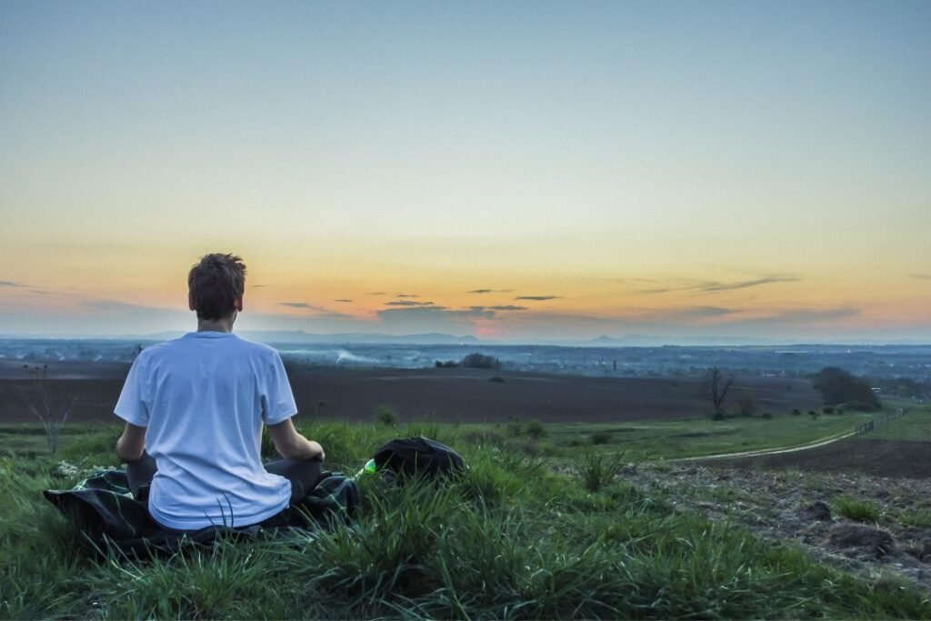 A peaceful outdoor scene of an adult sitting in lush green grass near the edge of a calm lake or ocean, quietly gazing toward the horizon. Soft golden-hour sunlight creates a warm, hopeful atmosphere, while the expansive water and open sky evoke a sense of reflection, possibility, and emotional healing. 